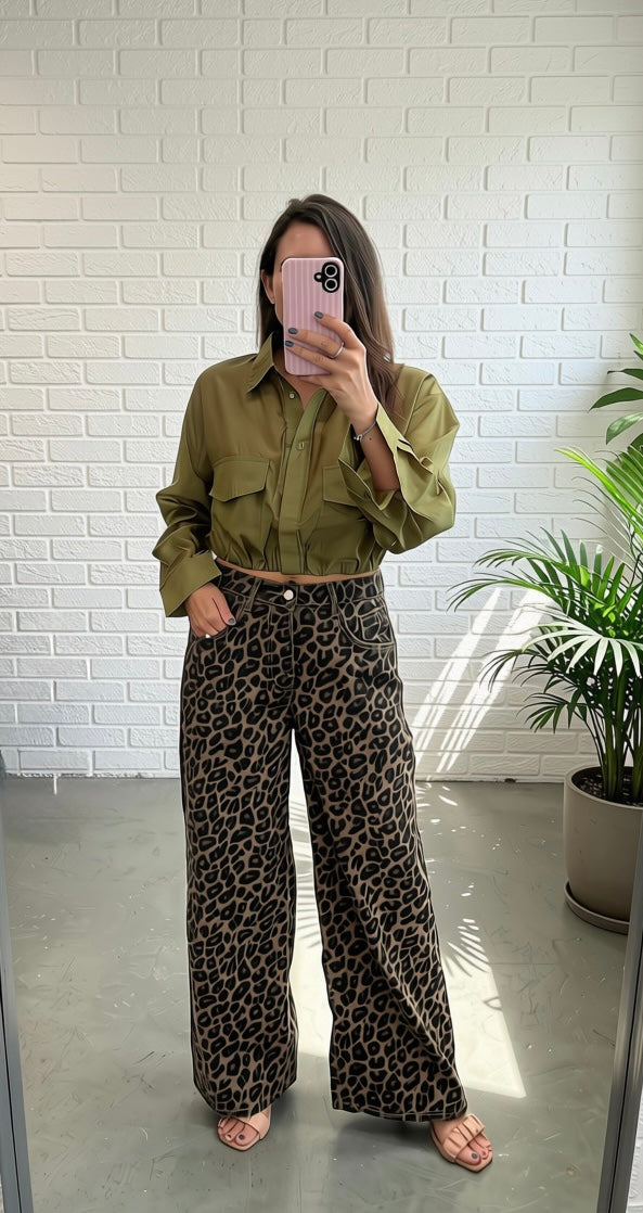 A woman takes a mirror selfie wearing an olive green cropped button-up shirt and high-waisted leopard-print wide-leg pants, standing in front of a white brick wall with a potted plant to the side.