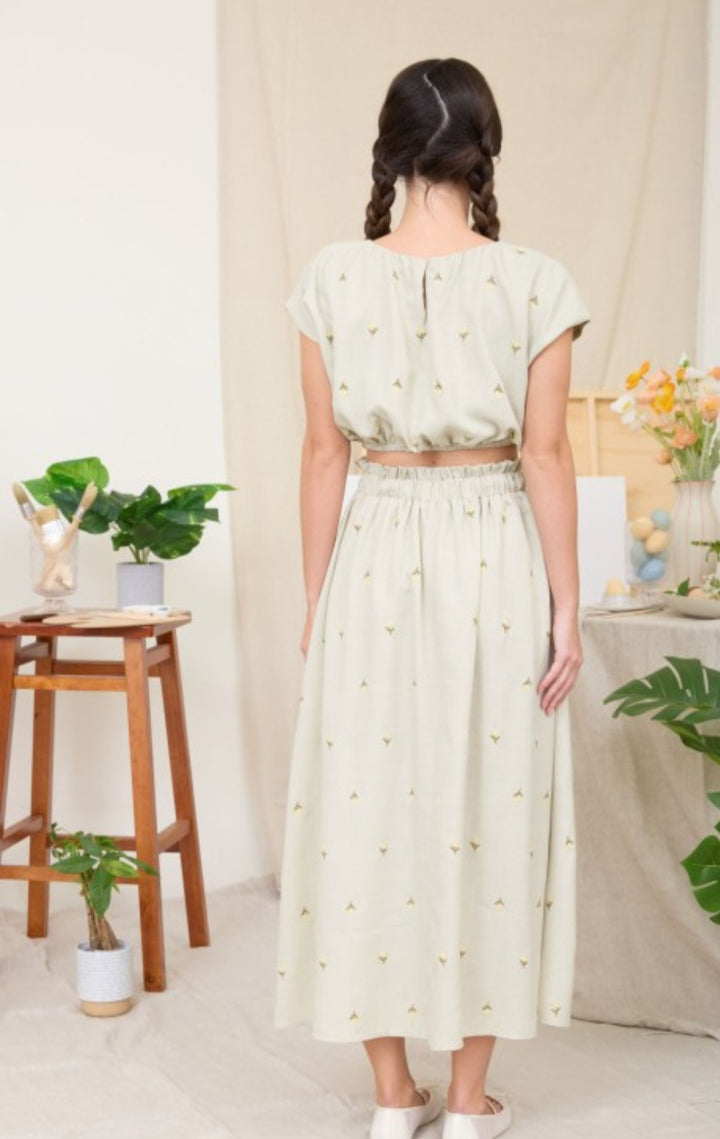Woman wearing a light green cropped skirt set with a matching top in a bright room with plants and furniture.