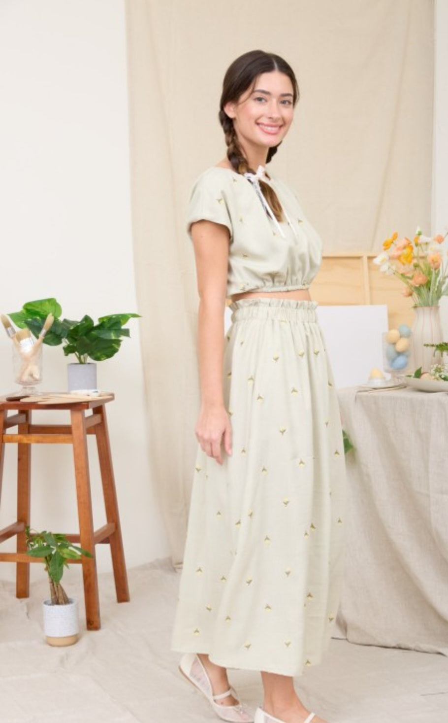 Woman in a light green outfit standing in a bright room with plants and a table.