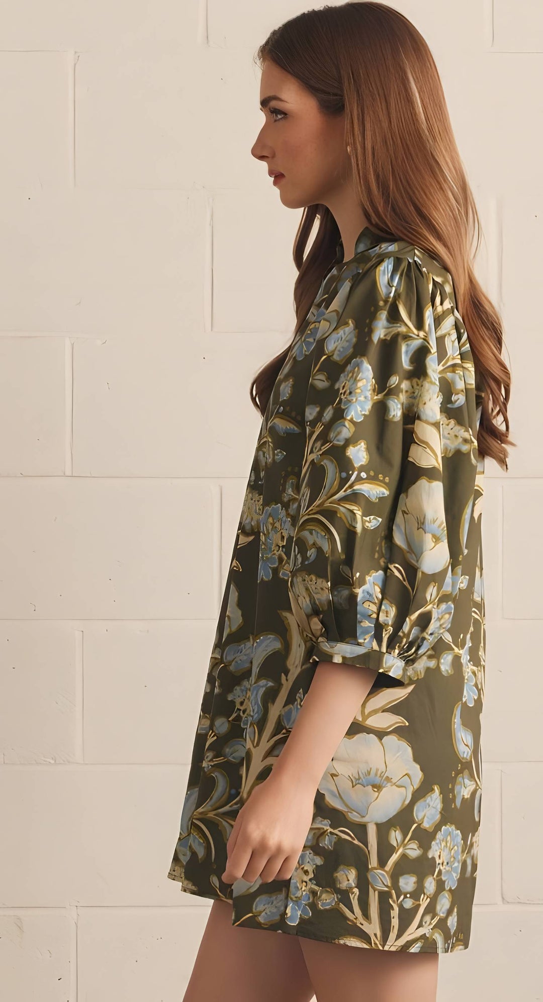 A woman in profile wearing a short-sleeve olive green floral dress with blue and beige flowers, standing against a white brick wall.
