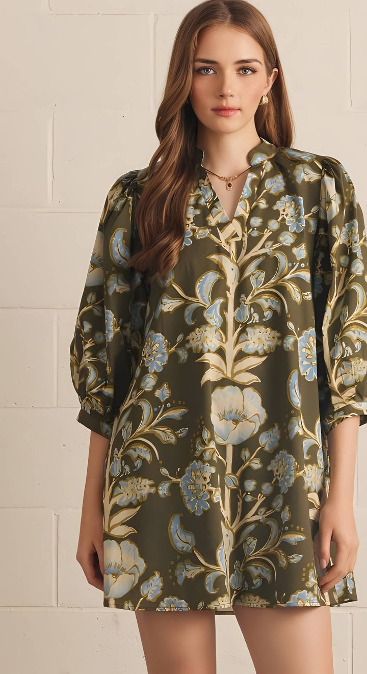 A woman modeling a short-sleeved, V-neck tunic dress with a green, beige, and blue floral pattern, standing against a white brick wall.