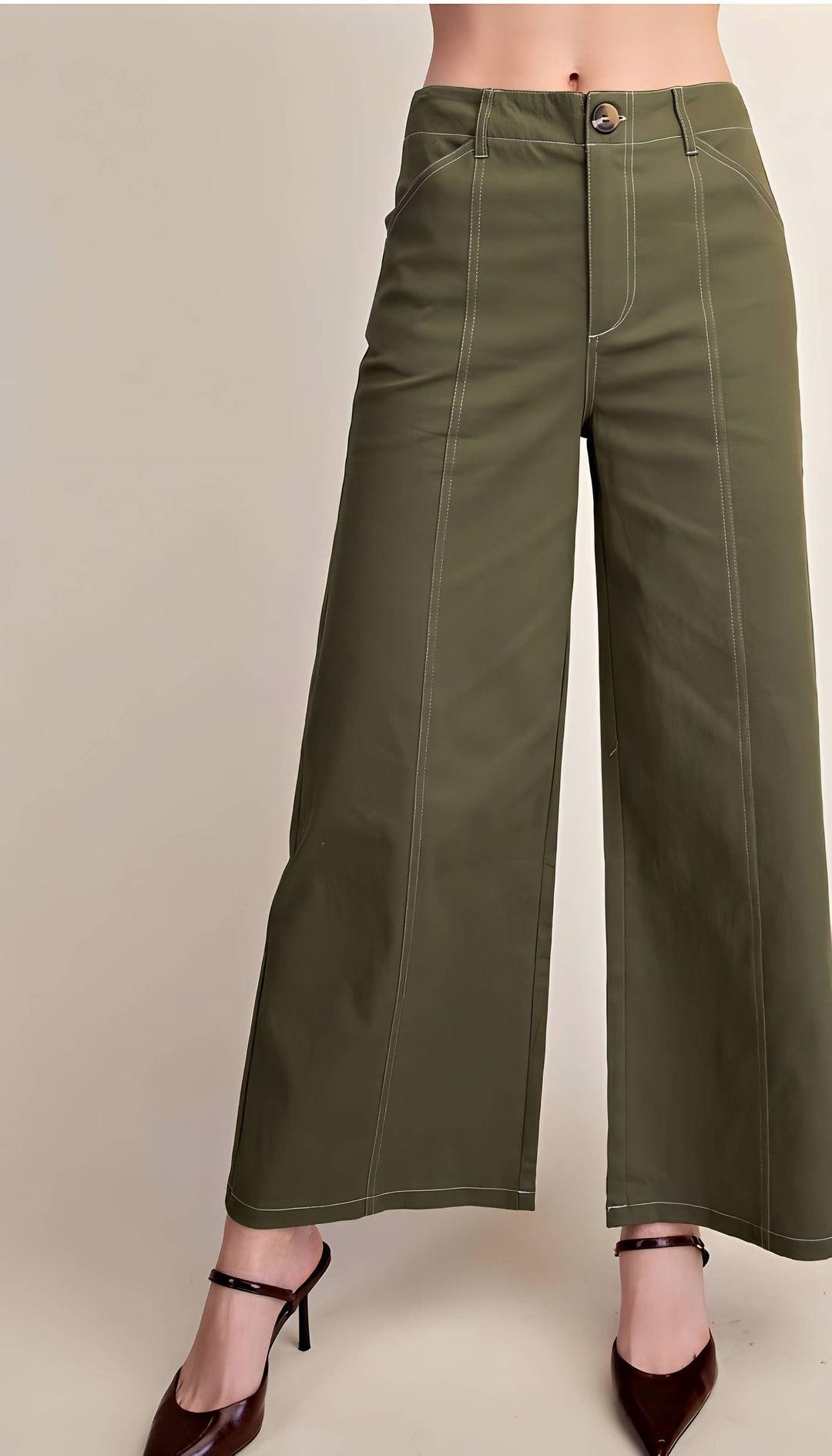 Olive green wide-leg cropped pants with contrast light stitching, shown on a model wearing dark brown pointed-toe heels against a plain beige studio background.