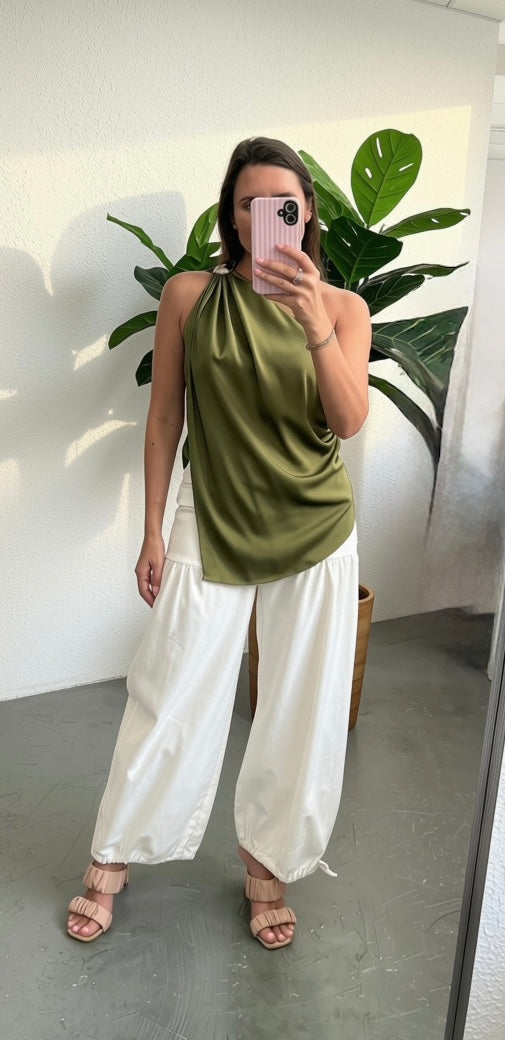A woman takes a mirror selfie wearing a sleeveless olive green satin halter top and white wide-leg pants, standing in a room with a large green plant in the background.