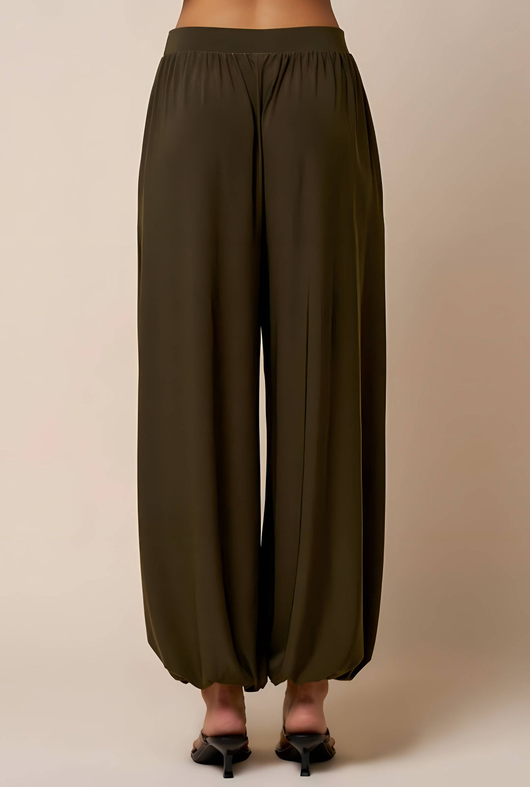 A person shown from the back wearing loose, wide-leg harem-style pants in an olive green color, paired with black pointed-toe mule heels against a plain beige studio background.