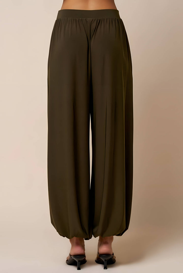 A person shown from the back wearing loose, wide-leg harem-style pants in an olive green color, paired with black pointed-toe mule heels against a plain beige studio background.