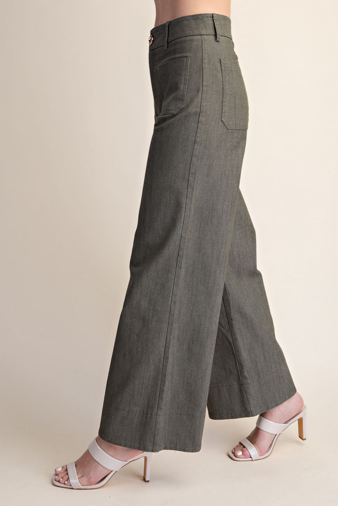 A person wearing high-waisted, wide-leg olive pants with side pockets, paired with white heeled sandals, shown from the waist down against a plain beige studio background.