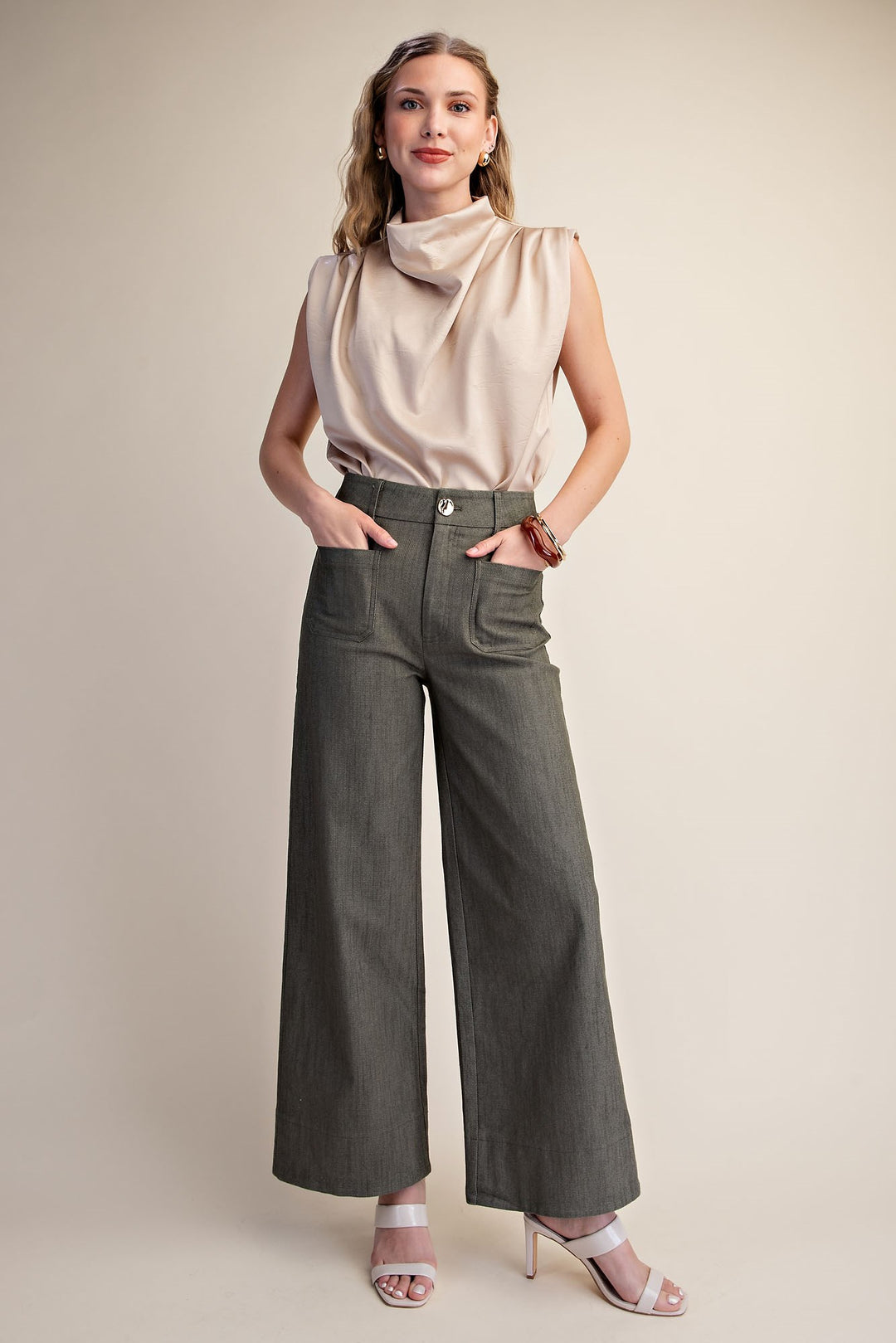 A woman modeling a sleeveless champagne beige cowl-neck top and dark olive green wide-leg pants, paired with light pink heeled sandals, against a solid beige studio background.