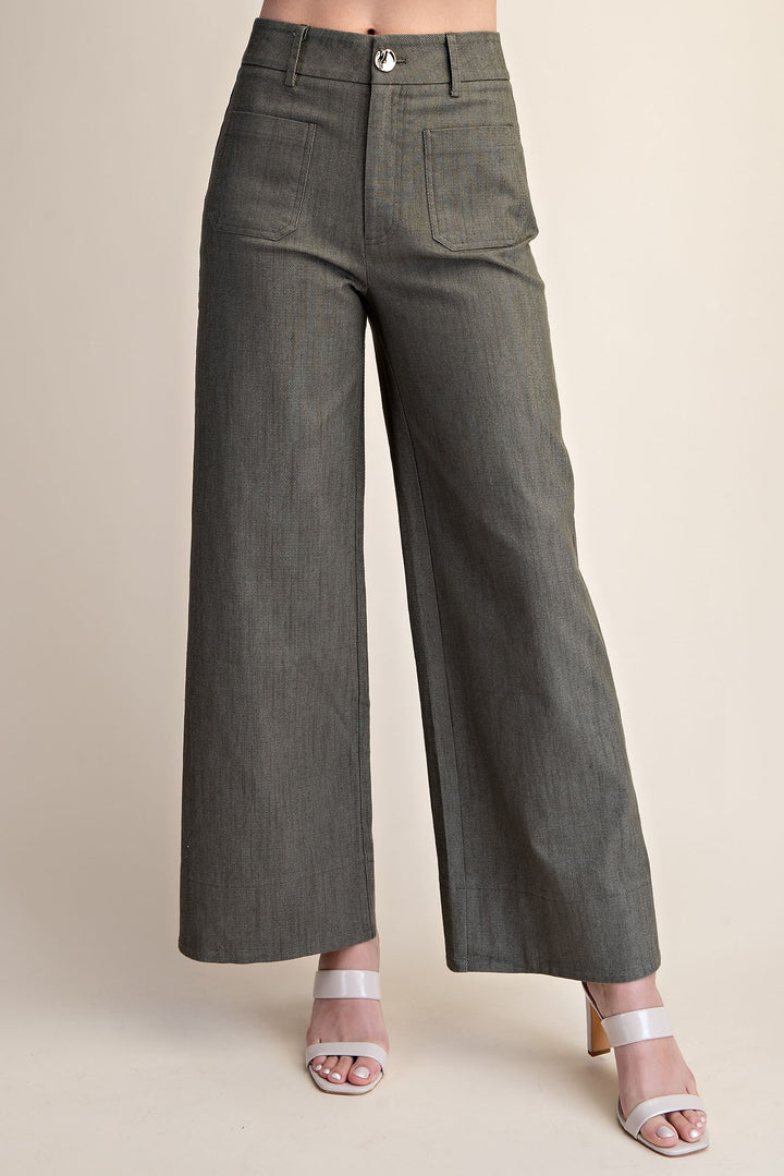 A person wearing high-waisted, wide-leg olive pants with front patch pockets and a button closure, paired with white heeled sandals against a plain beige studio background.