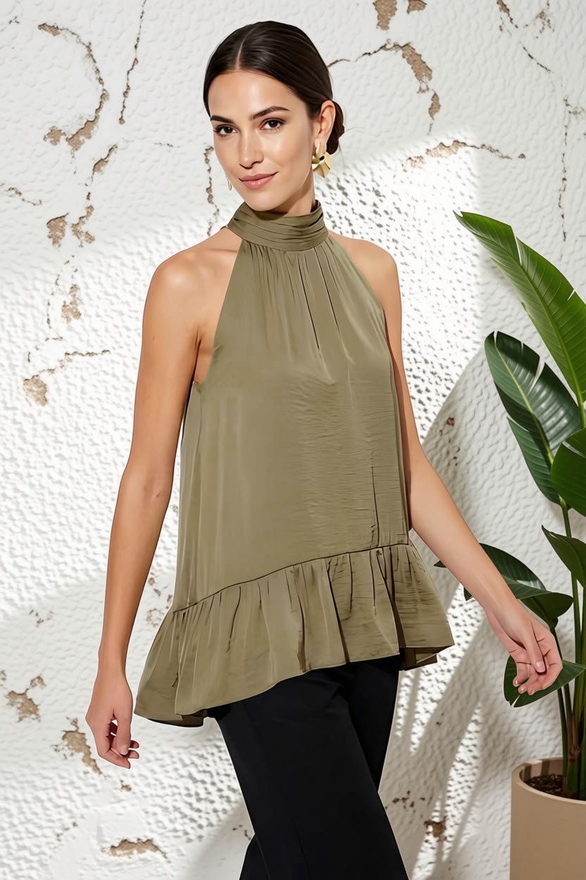 A woman modeling a sleeveless olive green halter top with a ruffled hem, paired with black pants, standing against a textured white wall with a green plant on the right.
