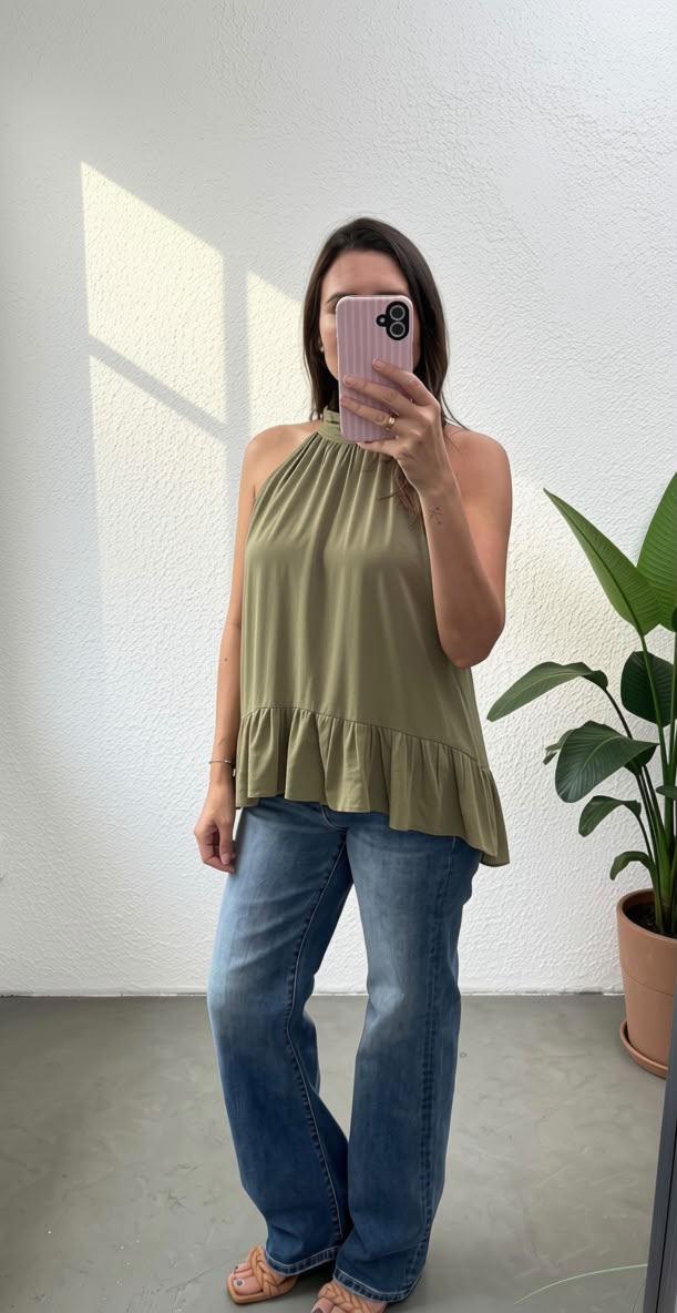 A woman takes a mirror selfie wearing a sleeveless olive green halter top with a ruffled hem, paired with blue jeans and pink sandals, standing in a brightly lit room with a potted plant.
