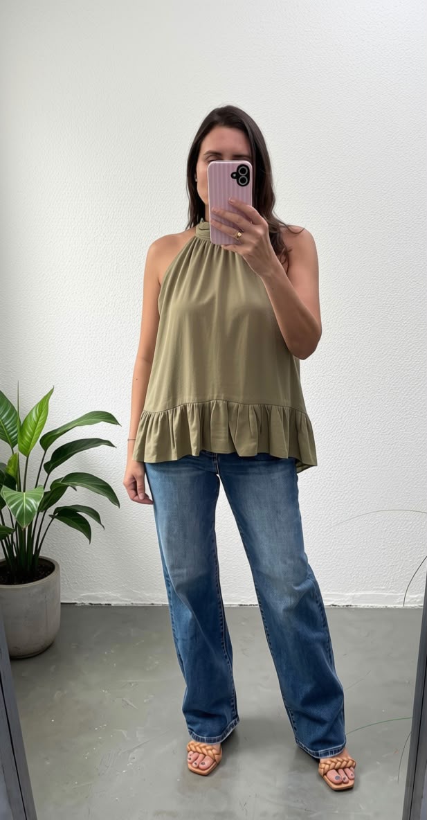A woman takes a mirror selfie wearing a sleeveless olive green halter top with a ruffled hem, paired with blue jeans and pink sandals, standing in a room with a white wall and a potted plant.