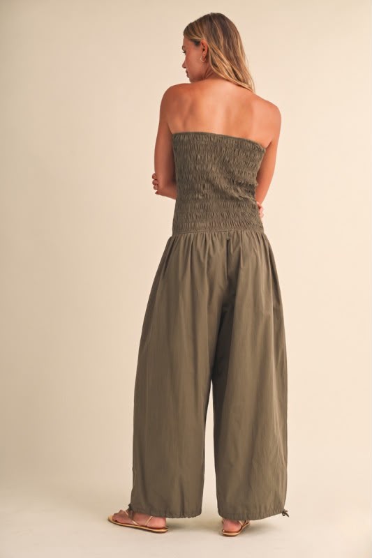 A woman shown from the back wearing a strapless smocked jumpsuit in an olive green color, paired with brown sandals, against a solid beige studio background.