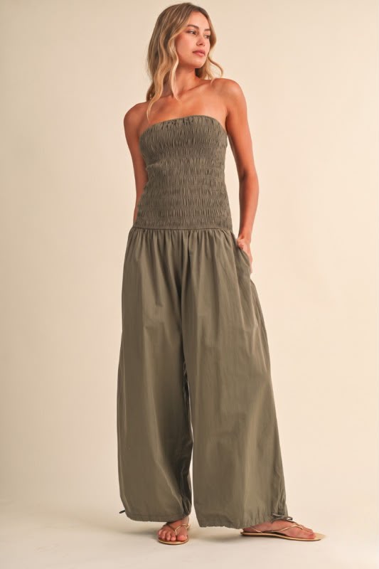 A woman modeling a strapless wide-leg jumpsuit in an olive green color, standing against a solid beige studio background.