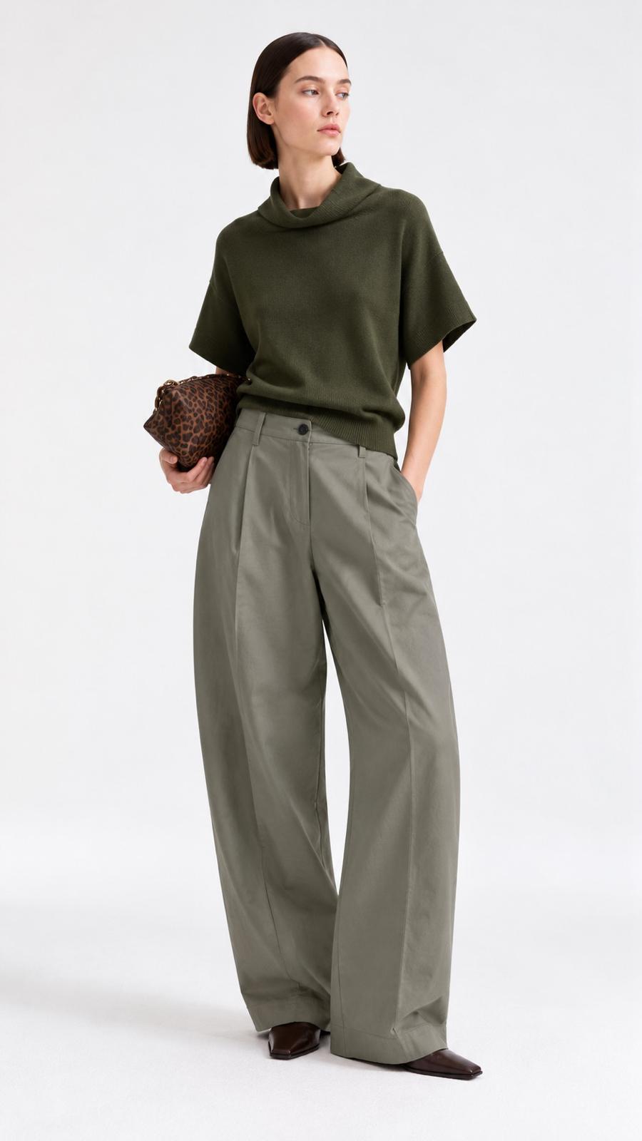 A model stands against a light gray studio background wearing an olive green short-sleeve mock-neck top and matching olive green wide-leg trousers, holding a brown textured clutch.