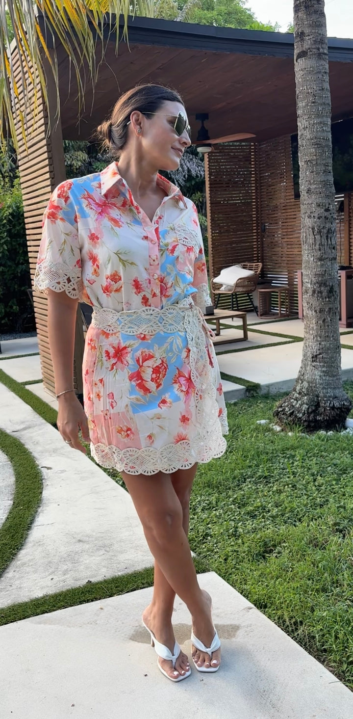 A woman wearing a short-sleeve floral print mini dress with lace trim and white flip-flops, standing outdoors on a patio near a house with palm trees.