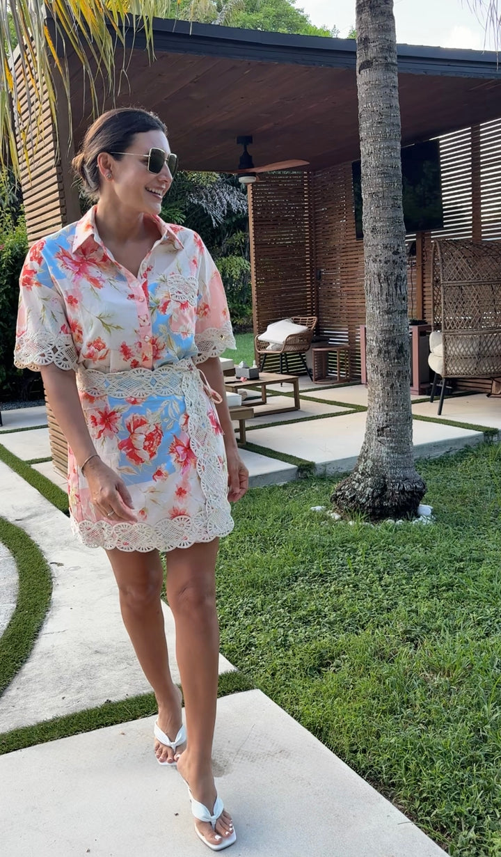 A woman wearing a short-sleeve floral mini shirt dress with lace trim walks outdoors on a patio near a tropical resort setting with palm trees and a wooden pavilion.