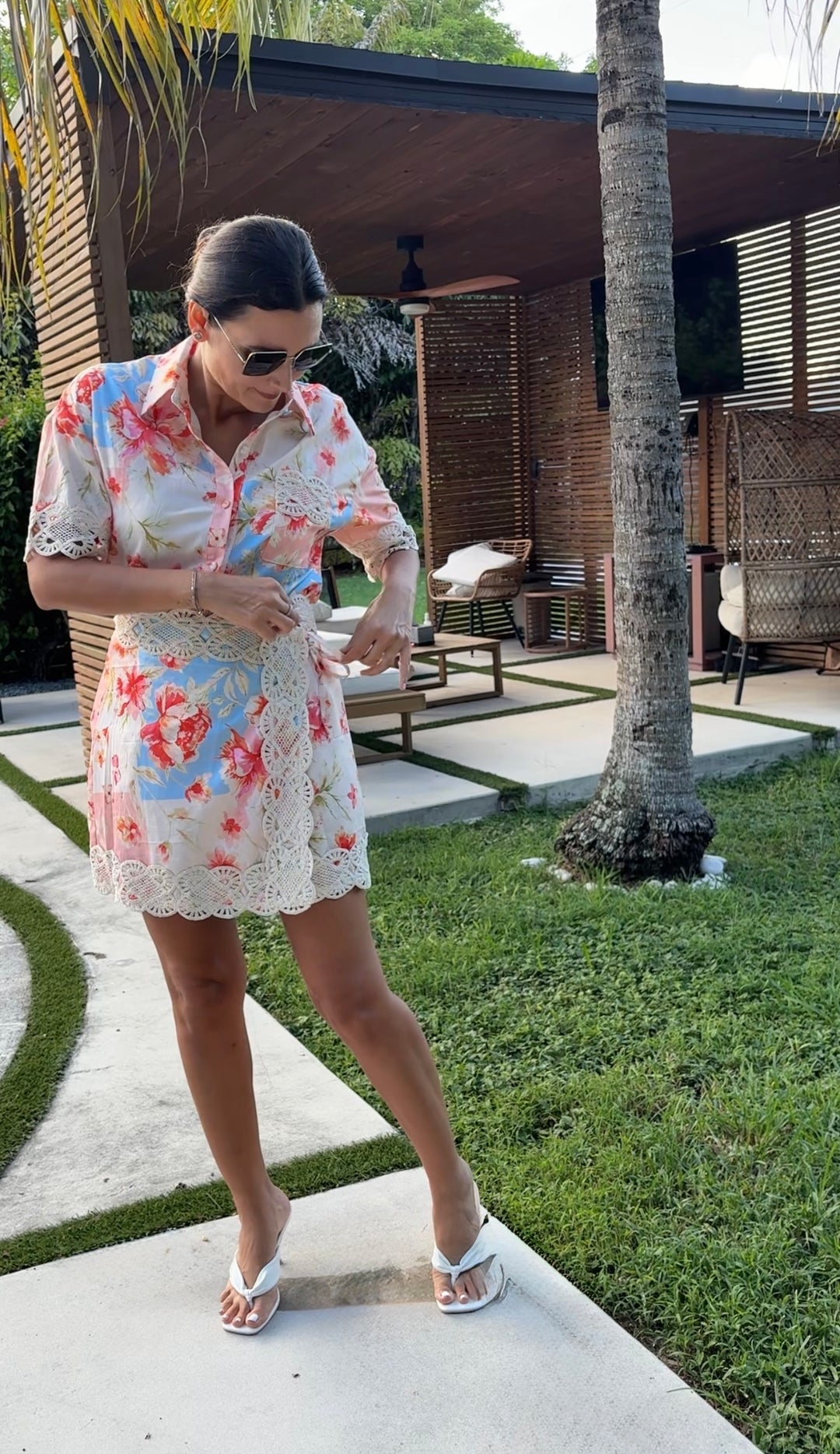 A woman wearing a short-sleeve floral lace mini dress and white sandals stands outdoors on a patio next to a palm tree, with a modern house and green lawn in the background.