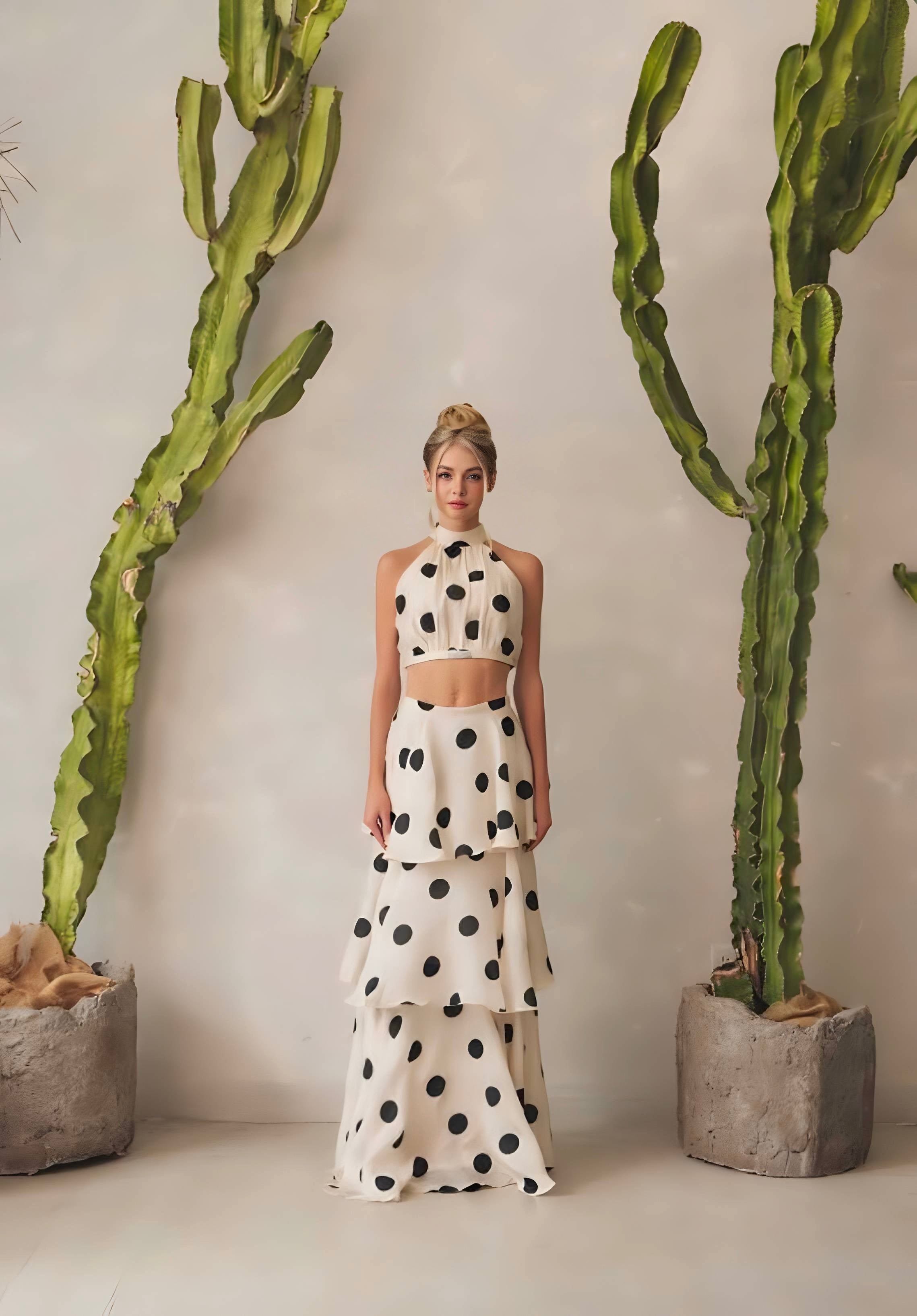 Woman in a polka dot outfit standing between two tall cacti indoors.