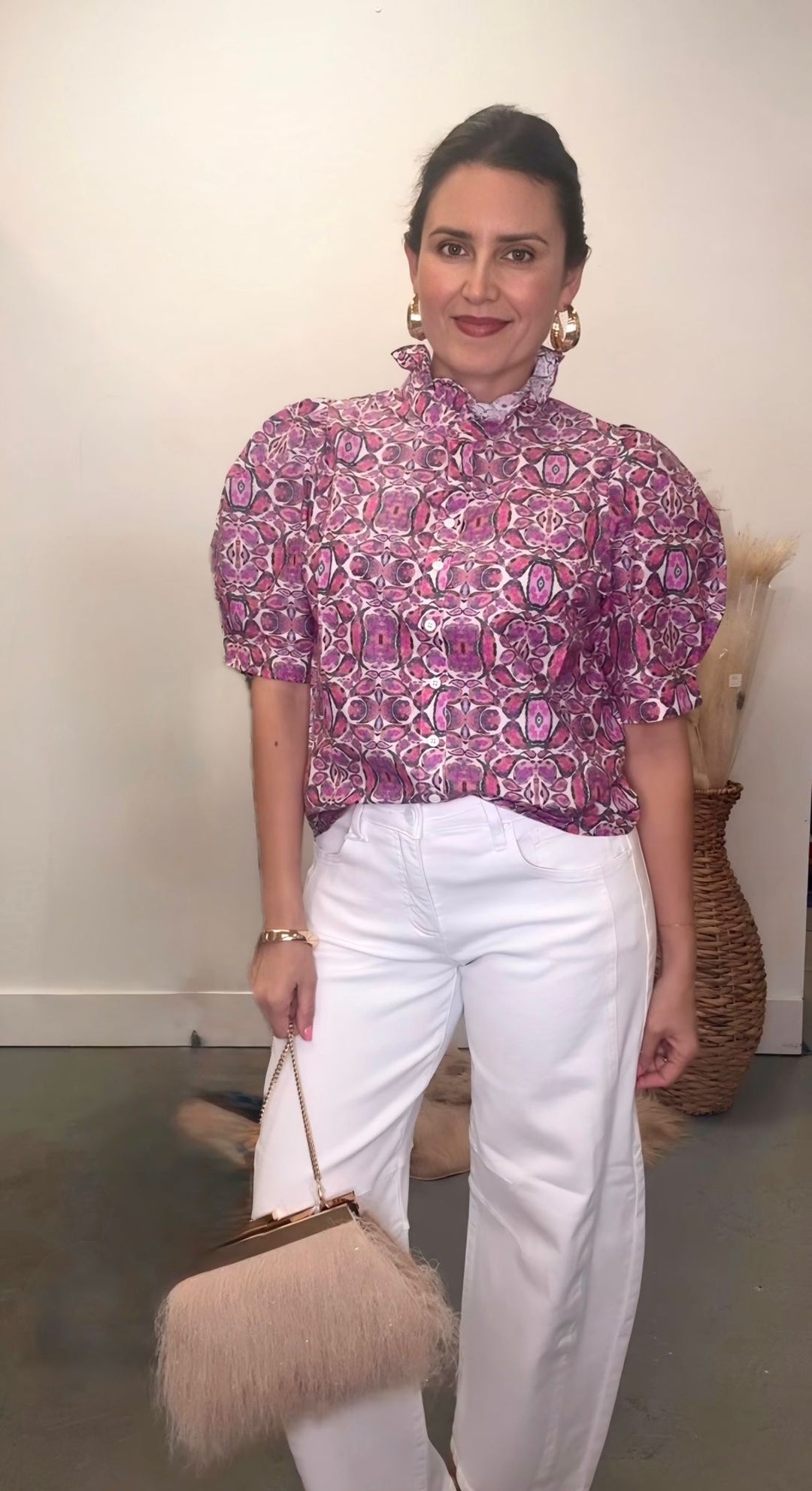 Woman wearing a patterned blouse and white pants, holding a beige handbag indoors.