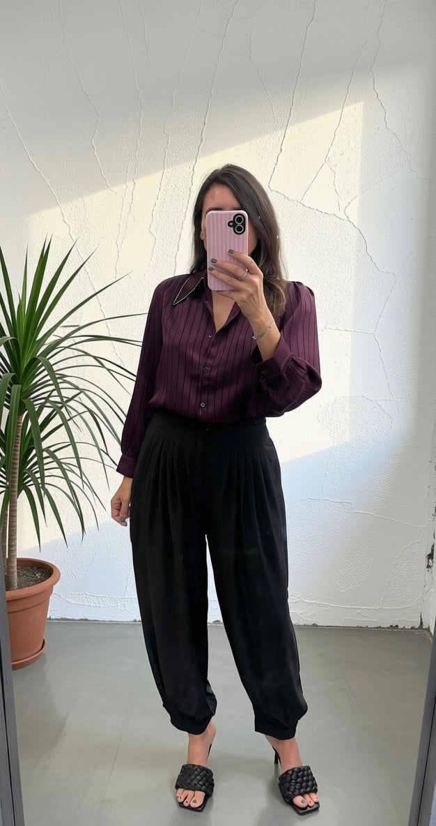 A woman takes a mirror selfie wearing a dark purple button-up shirt with black pinstripes and black wide-leg pants, standing in a room with a white wall and a potted plant.