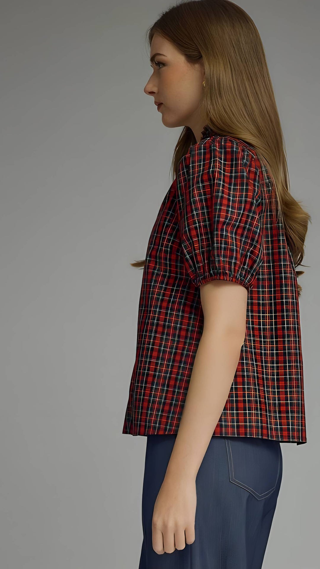 A woman shown in side profile wearing a short-sleeve puff-sleeve plaid shirt in red, black, and thin yellow lines, paired with navy blue pants, against a solid gray studio background.