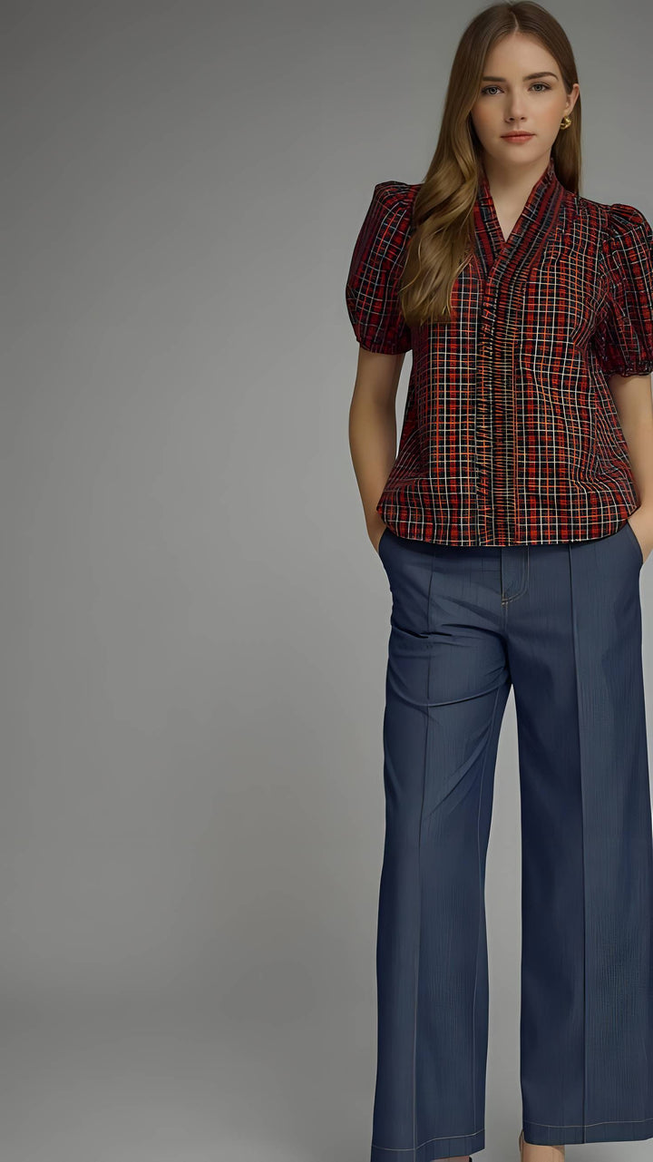 A woman modeling a short-sleeve plaid button-up blouse with puff sleeves and dark blue wide-leg trousers, photographed against a solid gray studio background.
