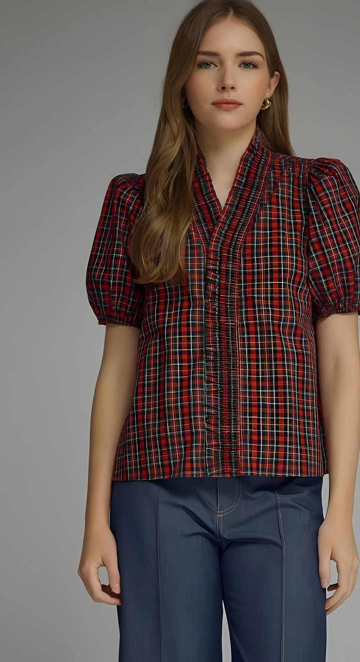 A woman modeling a short-sleeve plaid blouse with puff sleeves, featuring a red, black, and thin yellow/gold plaid pattern, paired with dark blue trousers against a solid gray studio background.