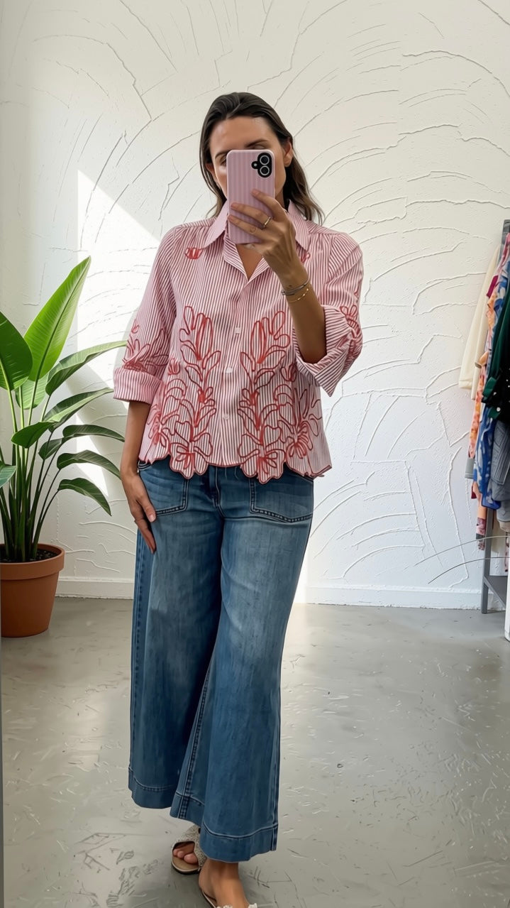 A woman takes a mirror selfie wearing a light pink and white striped button-up shirt with red floral embroidery on the front, paired with wide-leg blue jeans.