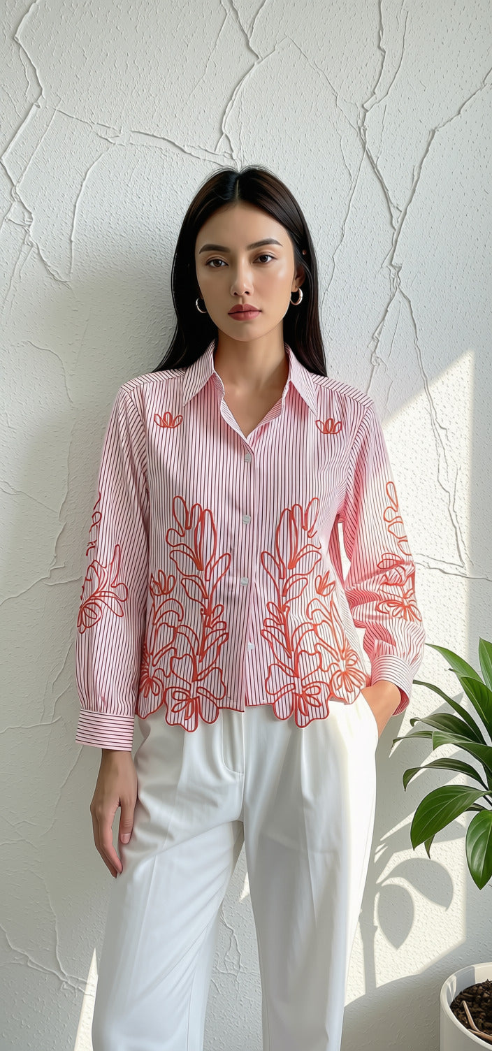 A woman stands against a textured white wall wearing a pink and white vertically striped long-sleeve button-up shirt with coral-red floral embroidery, paired with white trousers.