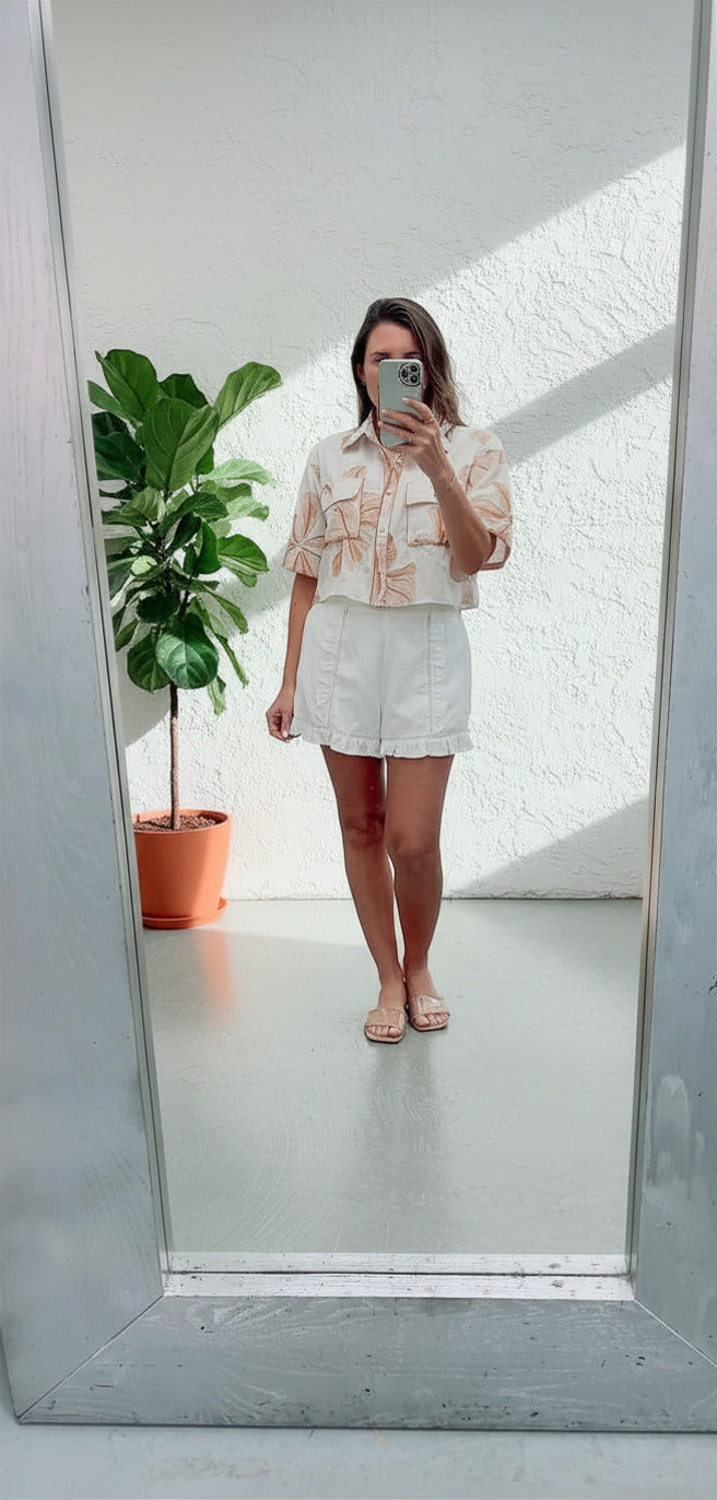 Person taking a mirror selfie wearing a light-colored outfit with a plant in the background.