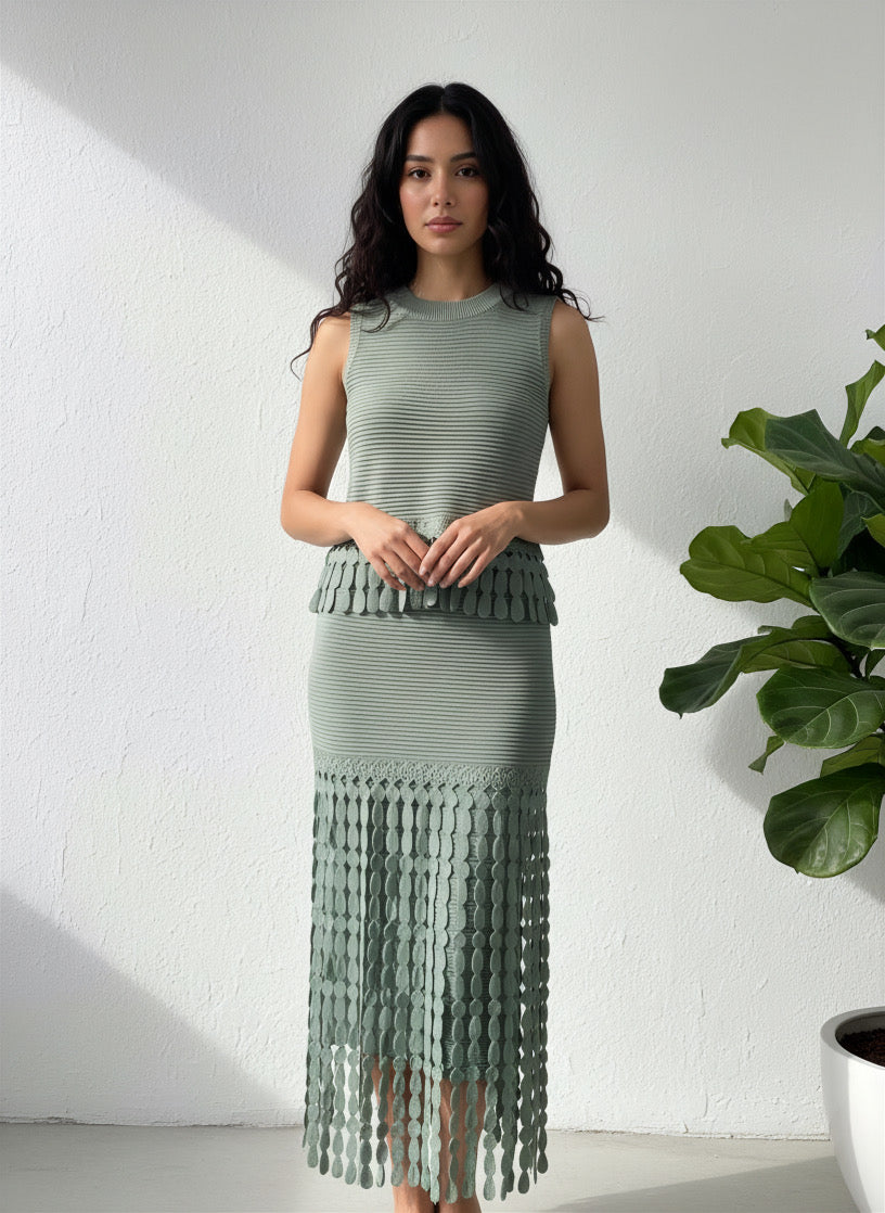 Woman wearing a green dress with a fringed skirt standing against a white wall with a plant to the side.