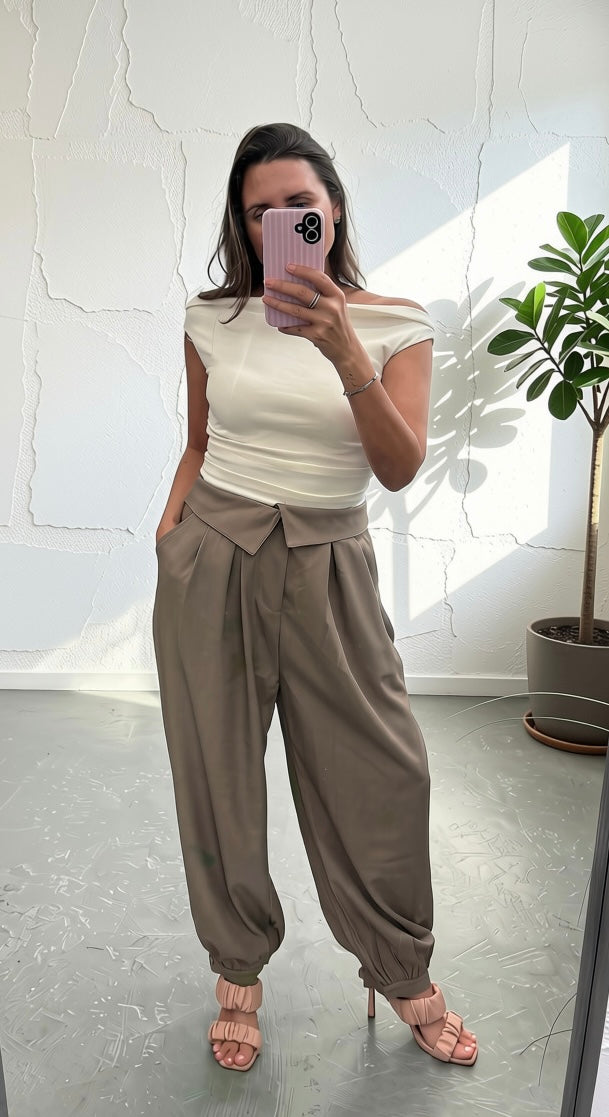 A woman takes a mirror selfie wearing a cream off-the-shoulder top and high-waisted, loose-fitting taupe-brown harem pants with elastic ankle cuffs, paired with pink heeled sandals.