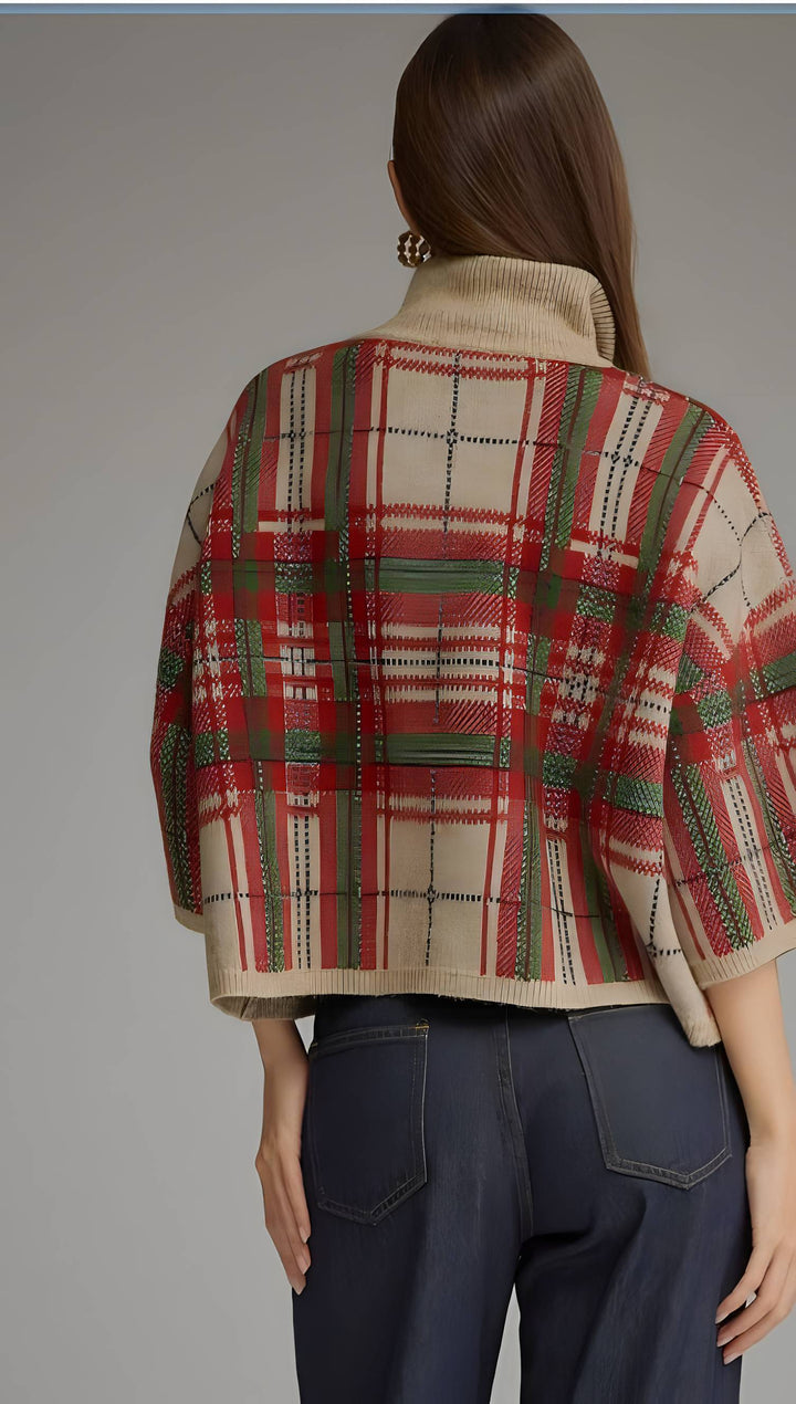 A woman shown from the back wearing a cropped knit cape cardigan with a red, green, and beige plaid pattern, paired with dark blue jeans, against a solid gray studio background.