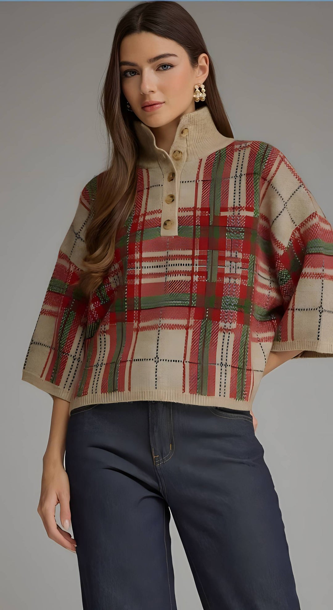 A woman modeling a cropped knit sweater with a high mock neck and wide sleeves, featuring a red, green, and beige plaid pattern with a buttoned placket. She is wearing dark blue jeans against a solid gray studio background.