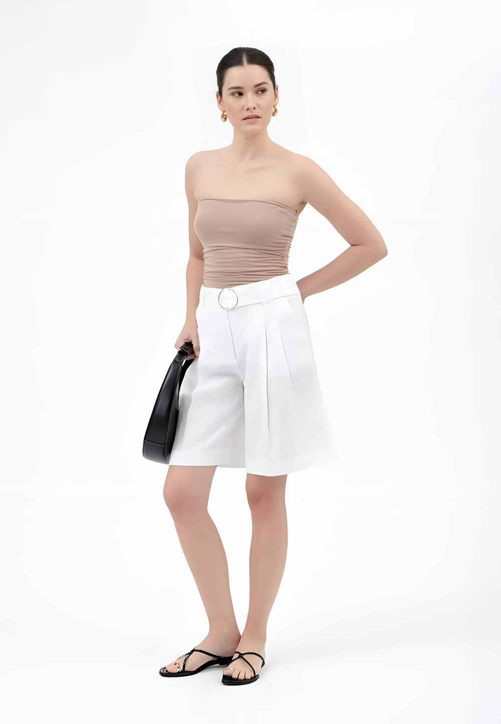 A model stands against a white studio background wearing a beige strapless top, white high-waisted pleated shorts with a white belt, and black flat sandals, while holding a black handbag.