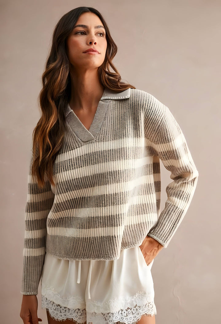 A woman wearing a ribbed knit long-sleeve sweater with horizontal gray and cream stripes, paired with white shorts, against a solid warm beige studio background.
