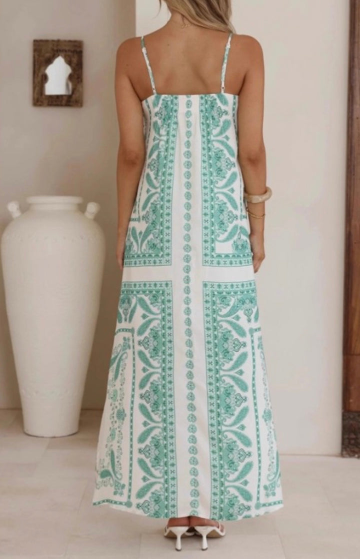 A woman viewed from behind wearing a sleeveless maxi dress with thin straps and a mint green and white bohemian-style floral pattern. She is standing indoors next to a large white ceramic vase.