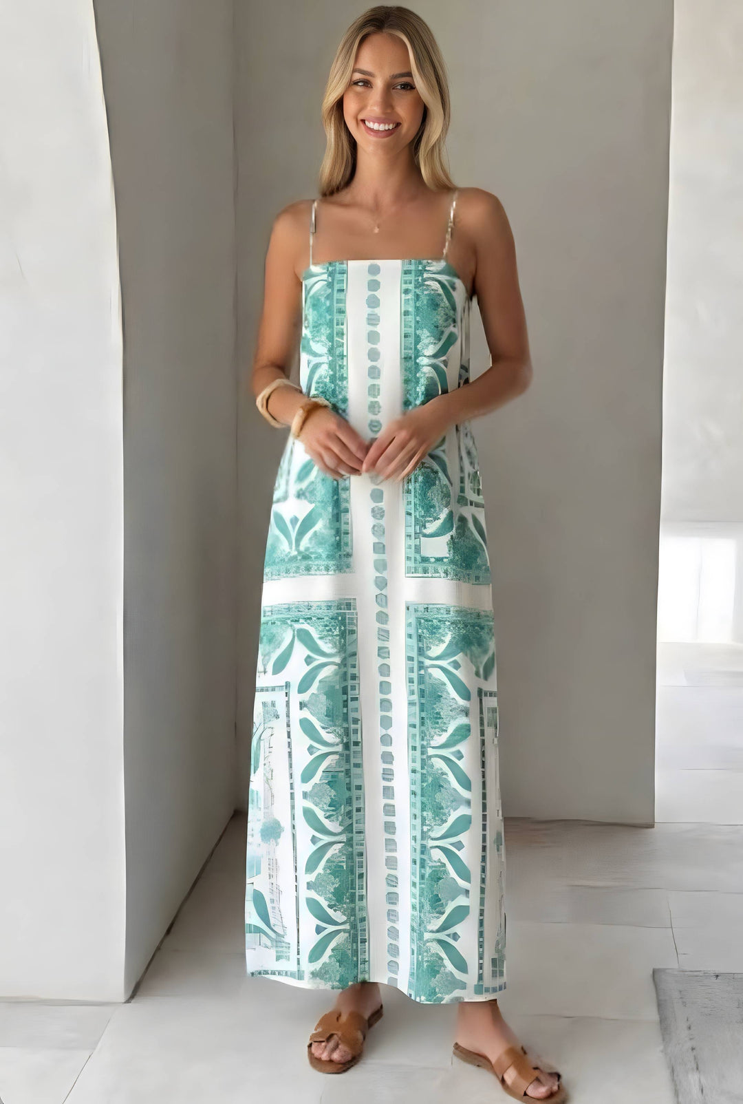 A woman modeling a sleeveless maxi dress with thin straps and a green and white botanical pattern, standing in a bright, modern indoor setting with white walls and tiled floors.