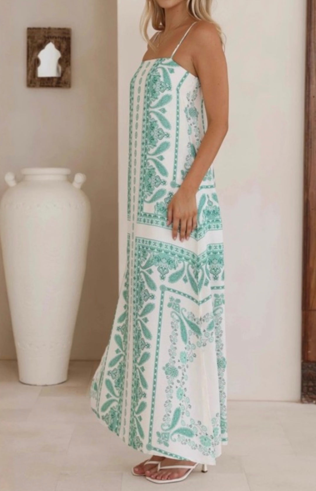 A woman wearing a white spaghetti-strap maxi dress with a mint green botanical pattern, standing in a brightly lit room next to a large white ceramic vase.