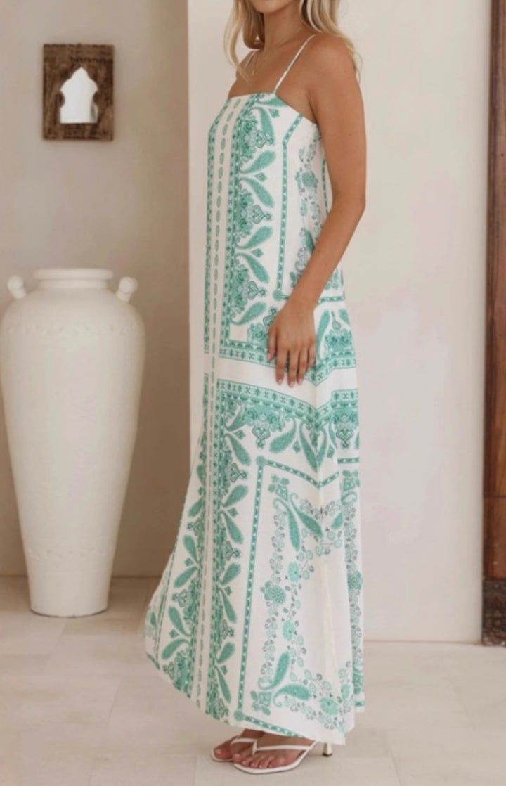A woman wearing a white spaghetti-strap maxi dress with a mint green botanical pattern, standing in a brightly lit room next to a large white ceramic vase.