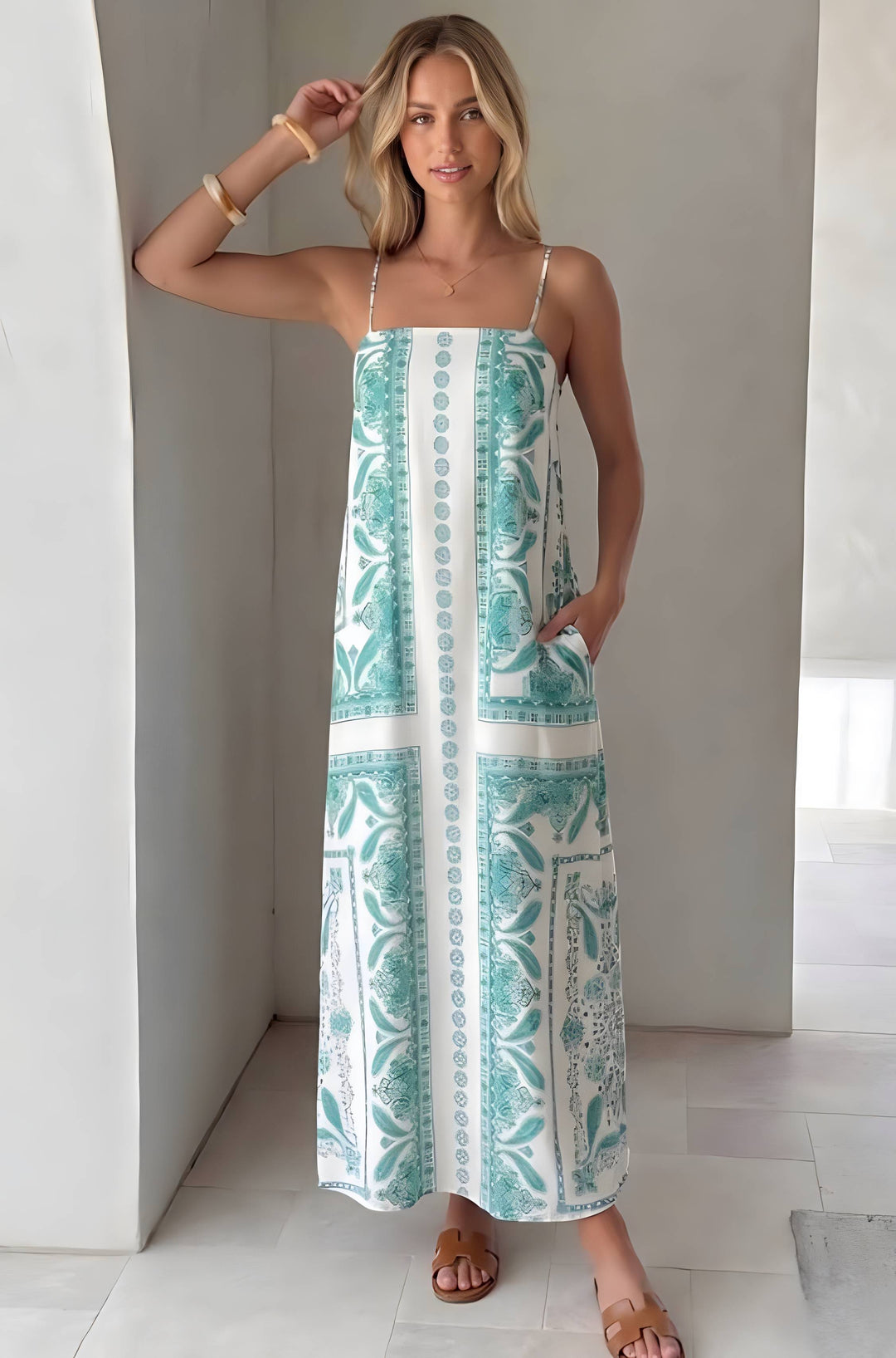 A woman modeling a sleeveless maxi dress with thin straps and a white base featuring a teal and green botanical pattern, standing in a bright, modern indoor setting.