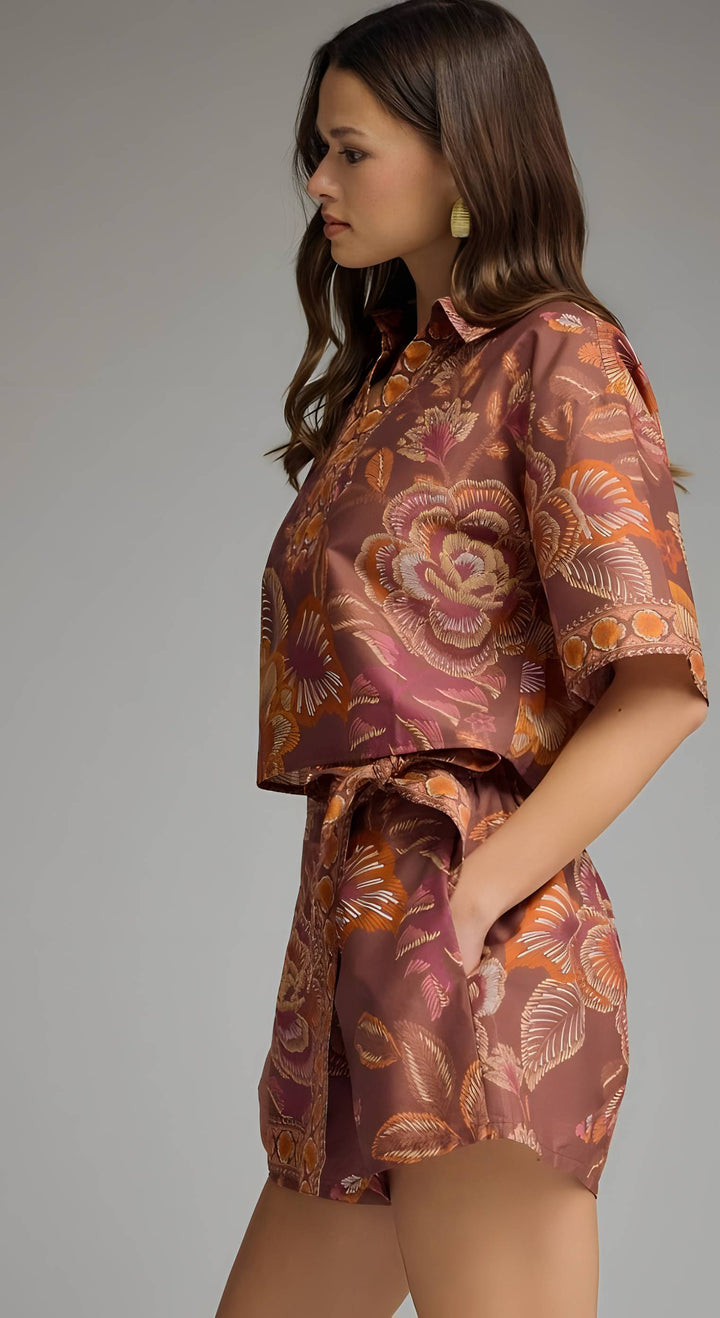 A woman wearing a short-sleeve collared shirt and matching shorts set in a rust-brown satin fabric with a large orange and beige floral pattern, photographed against a neutral gray studio background.