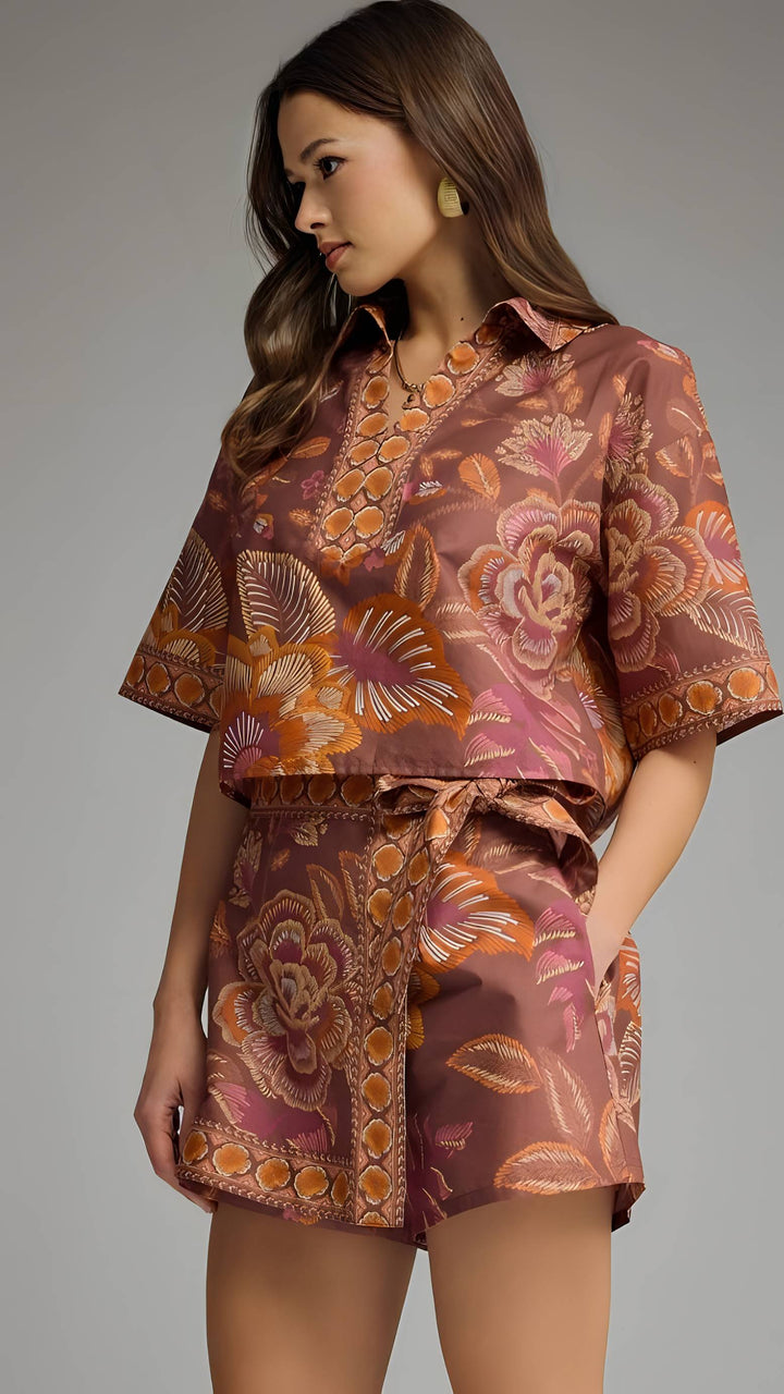 A woman modeling a matching short-sleeve button-front top and tie-waist shorts set in a brown and orange floral print with pink accents, photographed against a plain gray studio background.