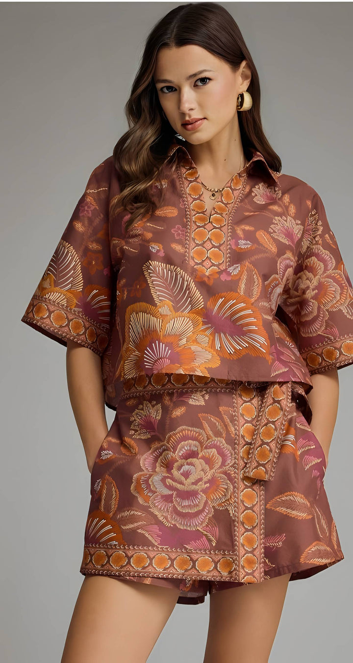 A woman modeling a matching two-piece set featuring a short-sleeve button-up shirt and coordinating shorts, both decorated with a brown, orange, and pink floral pattern.
