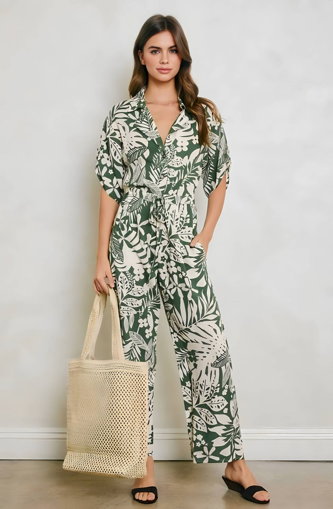 Woman wearing a green and white floral jumpsuit holding a woven bag against a plain background