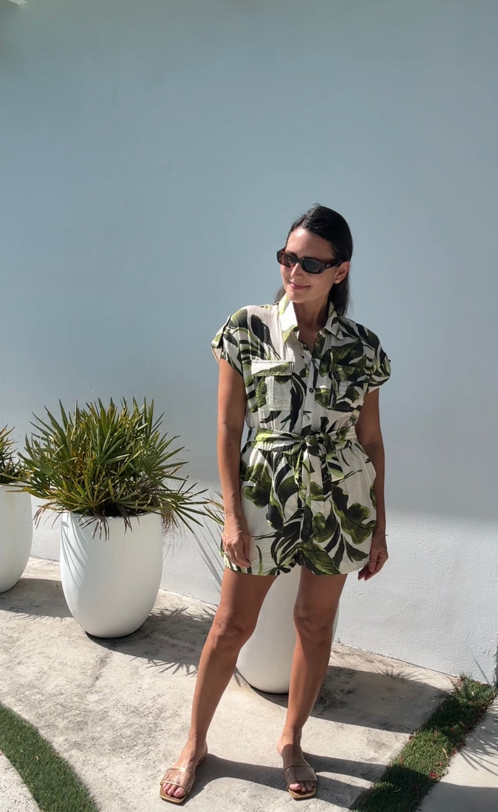 Woman wearing a tropical print romper standing outdoors with plants and a light blue wall in the background