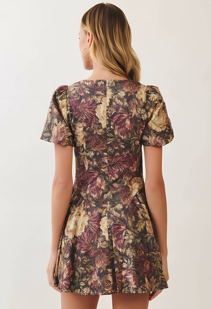 A woman viewed from the back wearing a short puff-sleeve dress with a dense floral print in burgundy, beige, and brown tones against a plain light background.