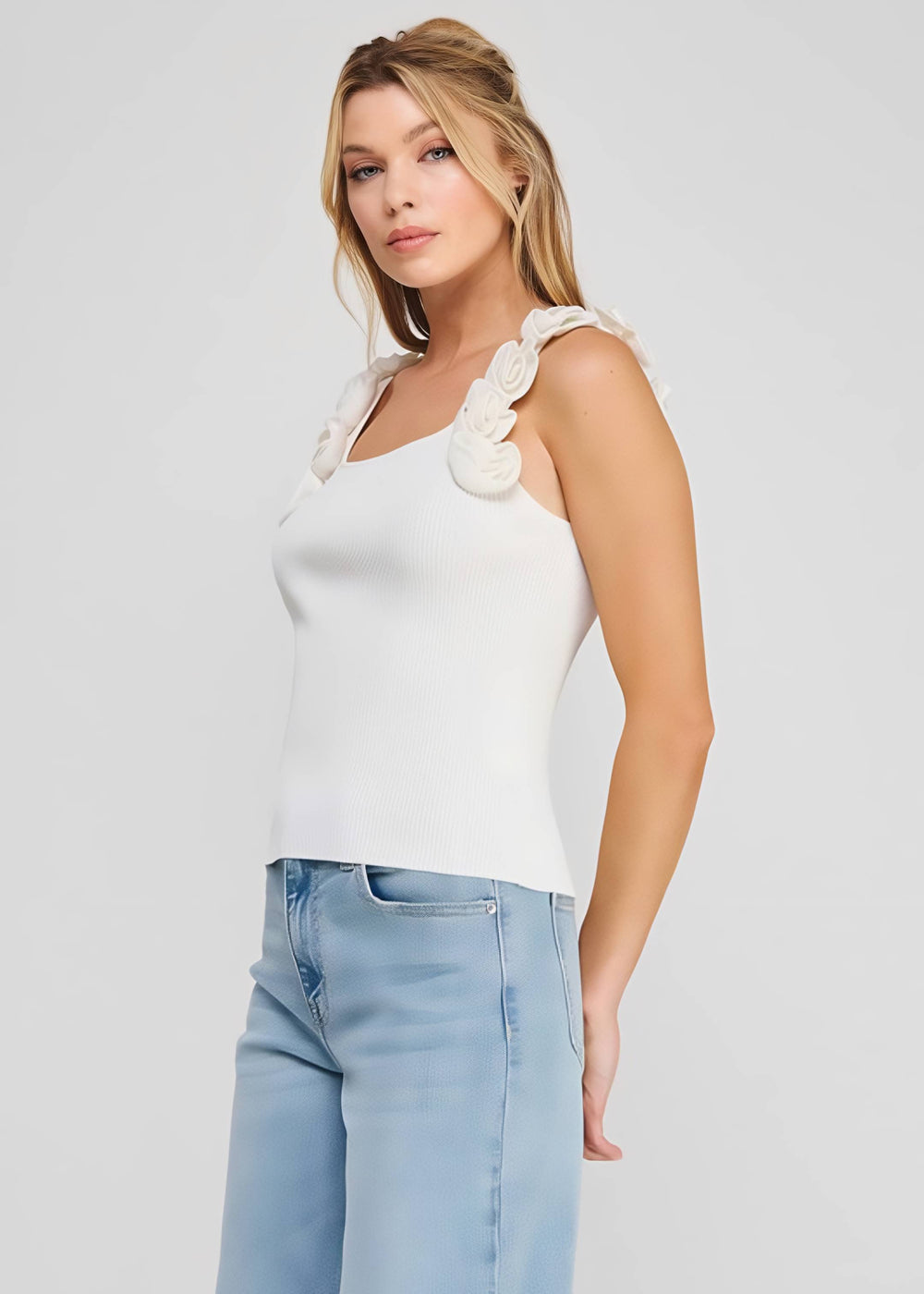 Woman wearing a white sleeveless top with floral details and light blue jeans on a plain background