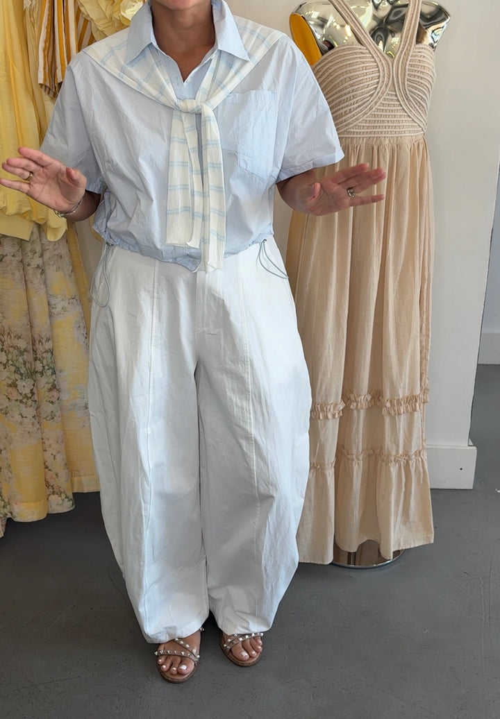 A person wearing a light blue short-sleeve button-up shirt with a cream-and-blue striped necktie, paired with loose white wide-leg pants and brown strappy sandals, standing indoors next to a beige dress on a mannequin.