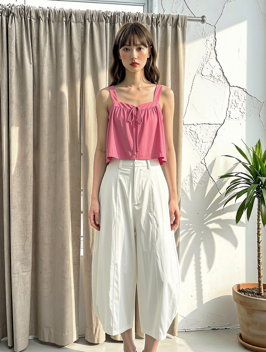 A woman stands indoors wearing a pink sleeveless ruffled top and white wide-leg cropped pants, posed in front of beige curtains and a white wall with a potted plant to the right.